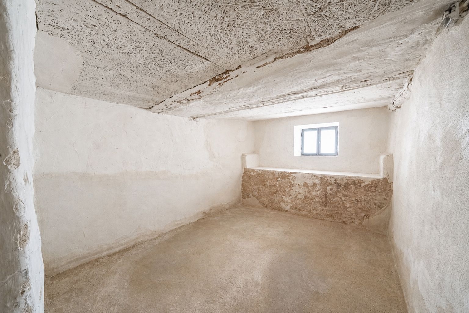 Leerer, weiß verputzter Kellerraum mit kleinem Fenster und rauer Decke.