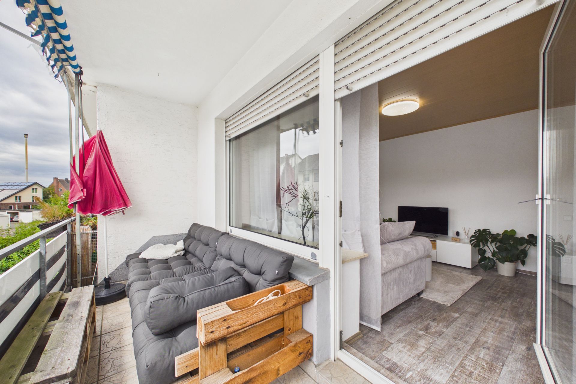 Balkon mit grauem Sofa, Markise und Blick ins Wohnzimmer mit Fernseher und Pflanzen.