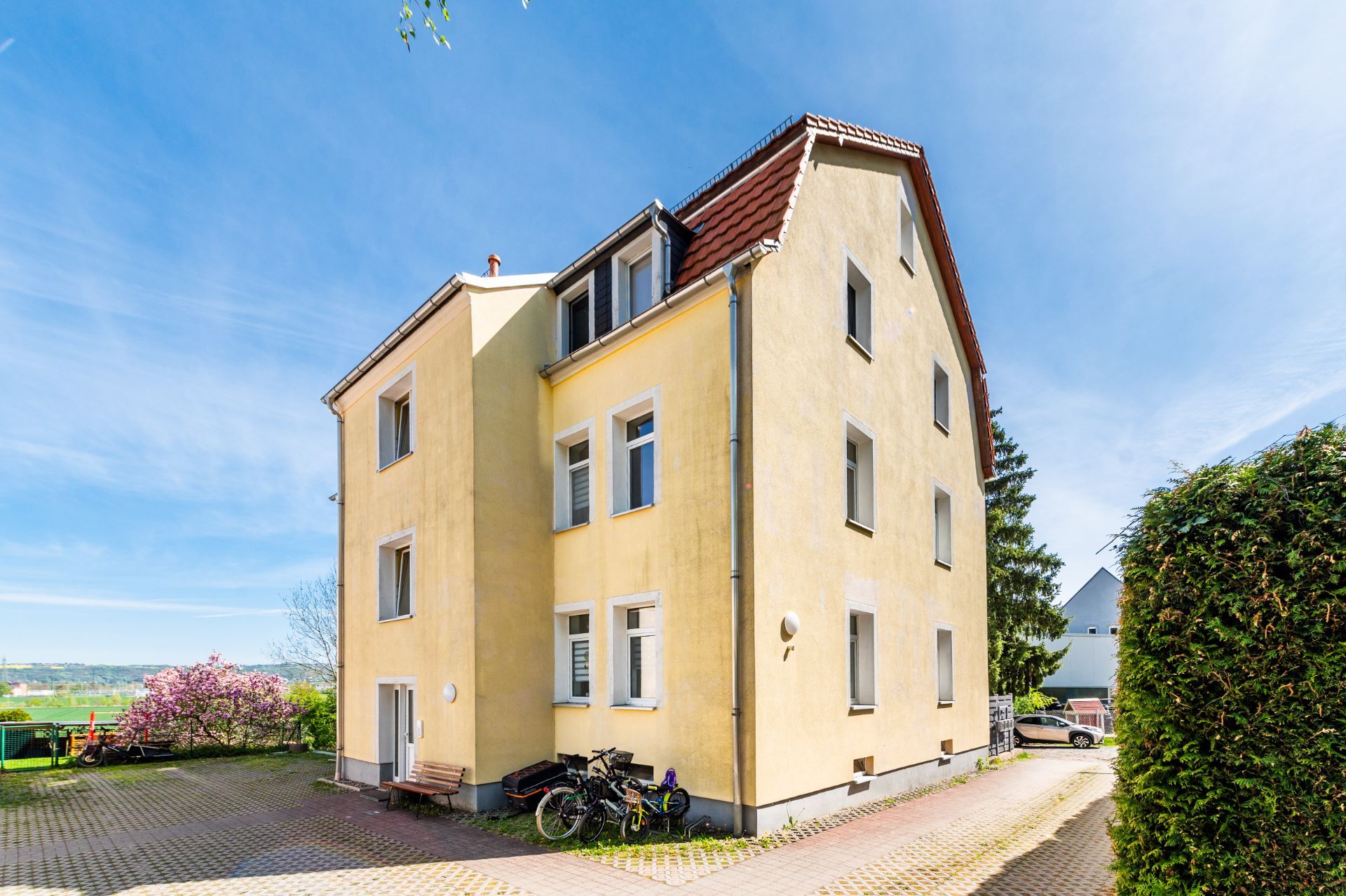 Gelbes Mehrfamilienhaus mit Satteldach, Fahrrädern und einer Bank davor auf gepflastertem Hof.