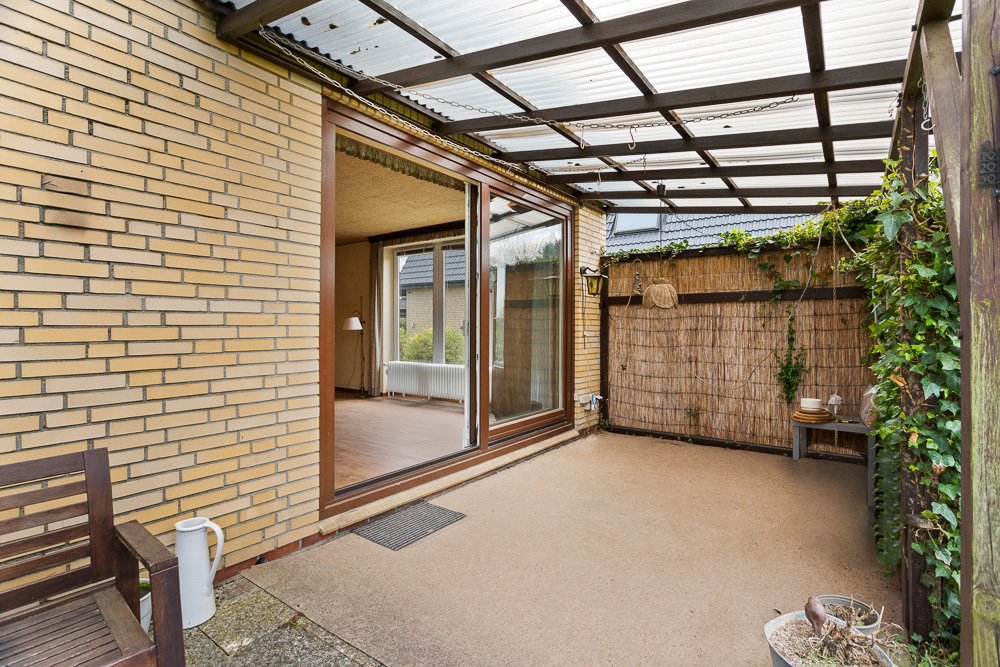 Überdachte Terrasse mit Holzmöbeln, offener Tür zum Wohnzimmer und Kletterpflanzen an den Wänden.