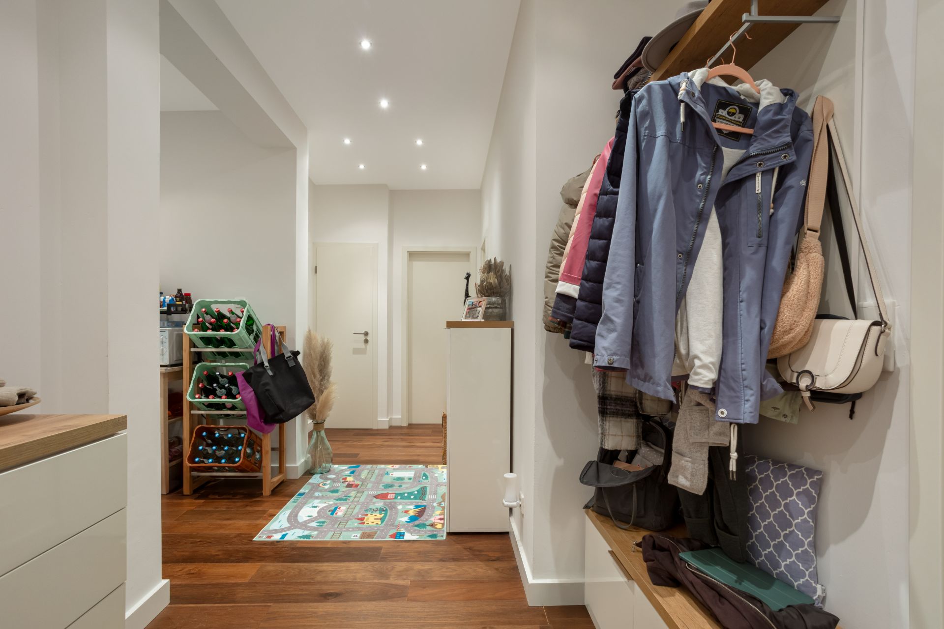 Flur mit Holzfußboden, Garderobe mit Jacken, Sitzbank und Teppich mit buntem Muster.