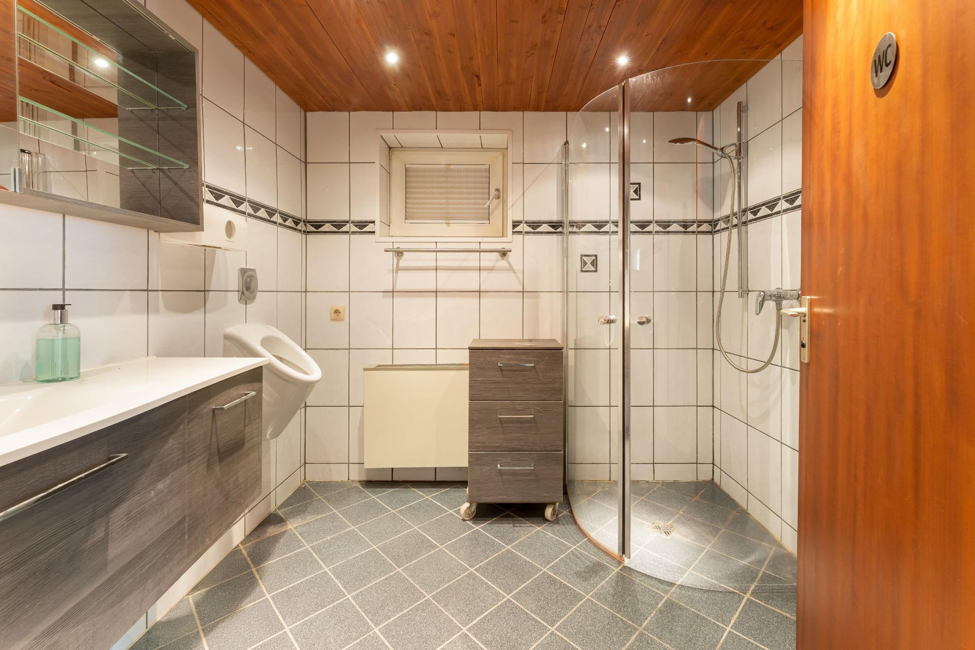 Helles, gefliestes Badezimmer mit Dusche, Waschbecken, Urinal und Holzdecke.