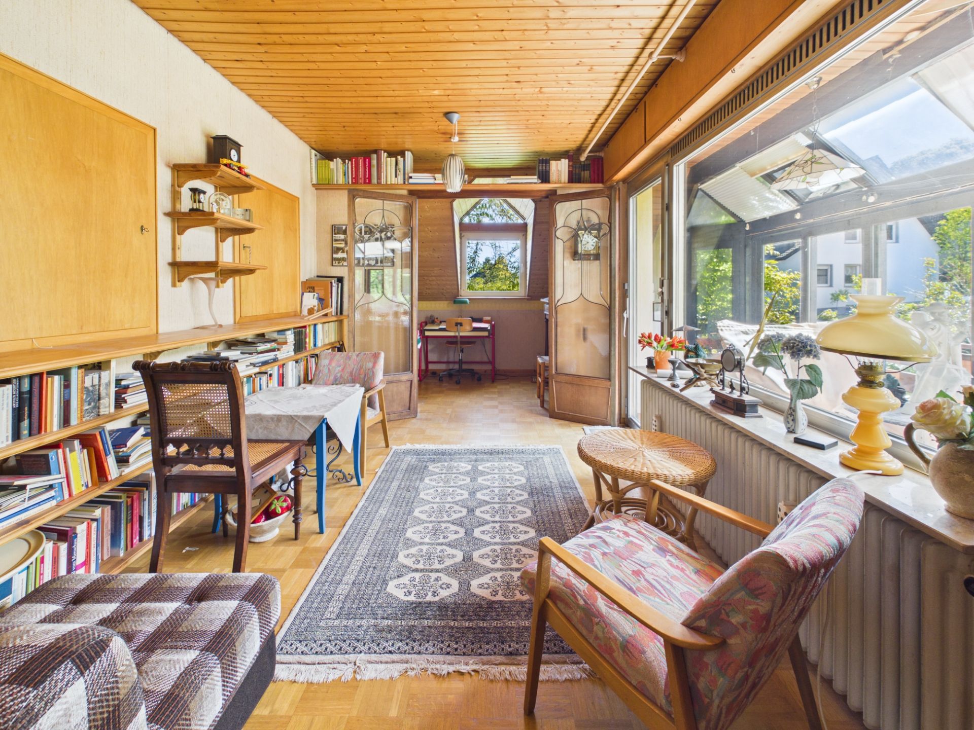 Helles Wohnzimmer mit großen Fenstern, Teppich, Regalen, Sessel und Schreibtisch im Hintergrund.