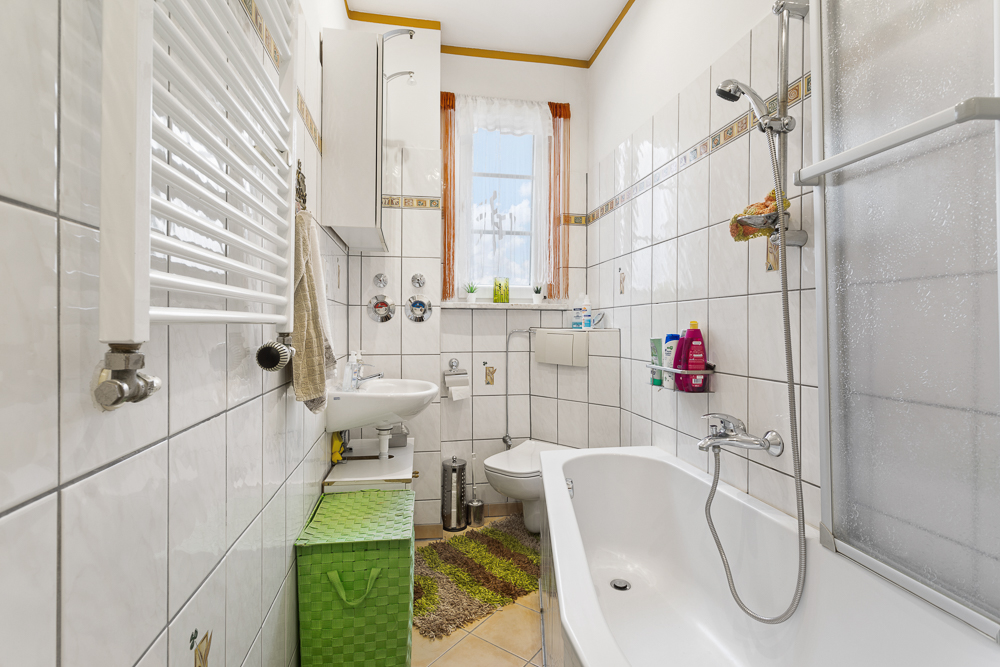 Helles Badezimmer mit Badewanne, Waschbecken, Bidet und Fenstern, weiße Fliesen und bunte Accessoires.