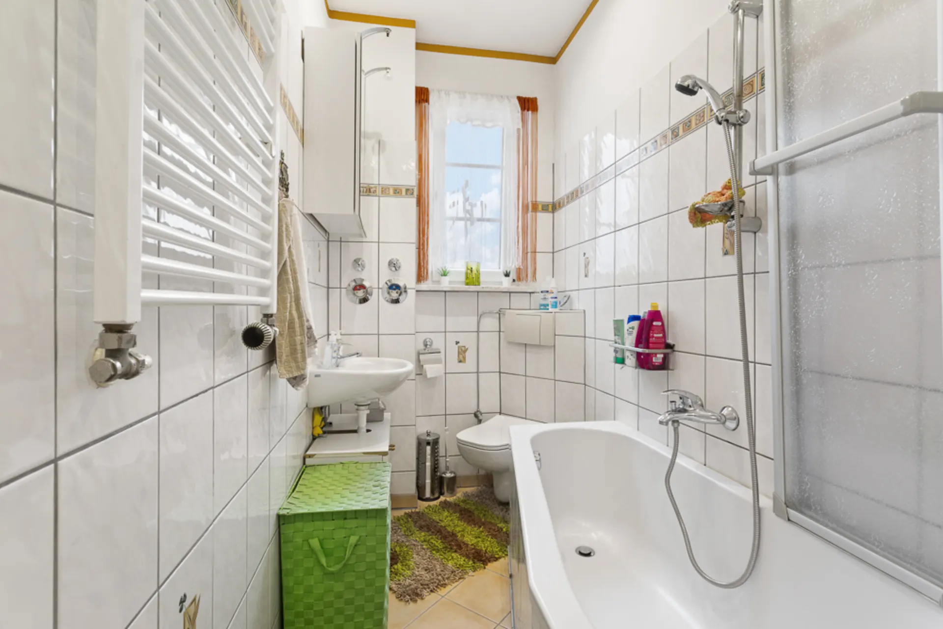 Helles Badezimmer mit Badewanne, Waschbecken, Bidet und Fenstern, weiße Fliesen und bunte Accessoires.