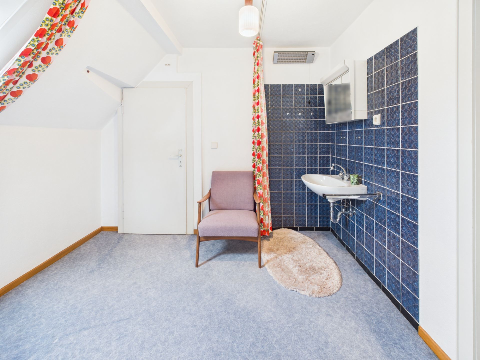 Helles Zimmer mit Sessel, Waschbecken, blauen Fliesen und buntem Vorhang.