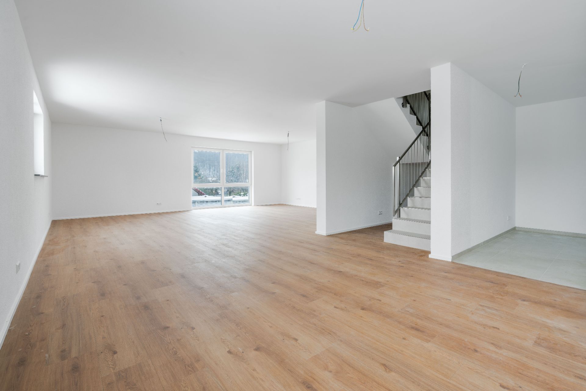 Helles, leeres Wohnzimmer mit Holzfußboden und Treppe, große Fenster im Hintergrund.