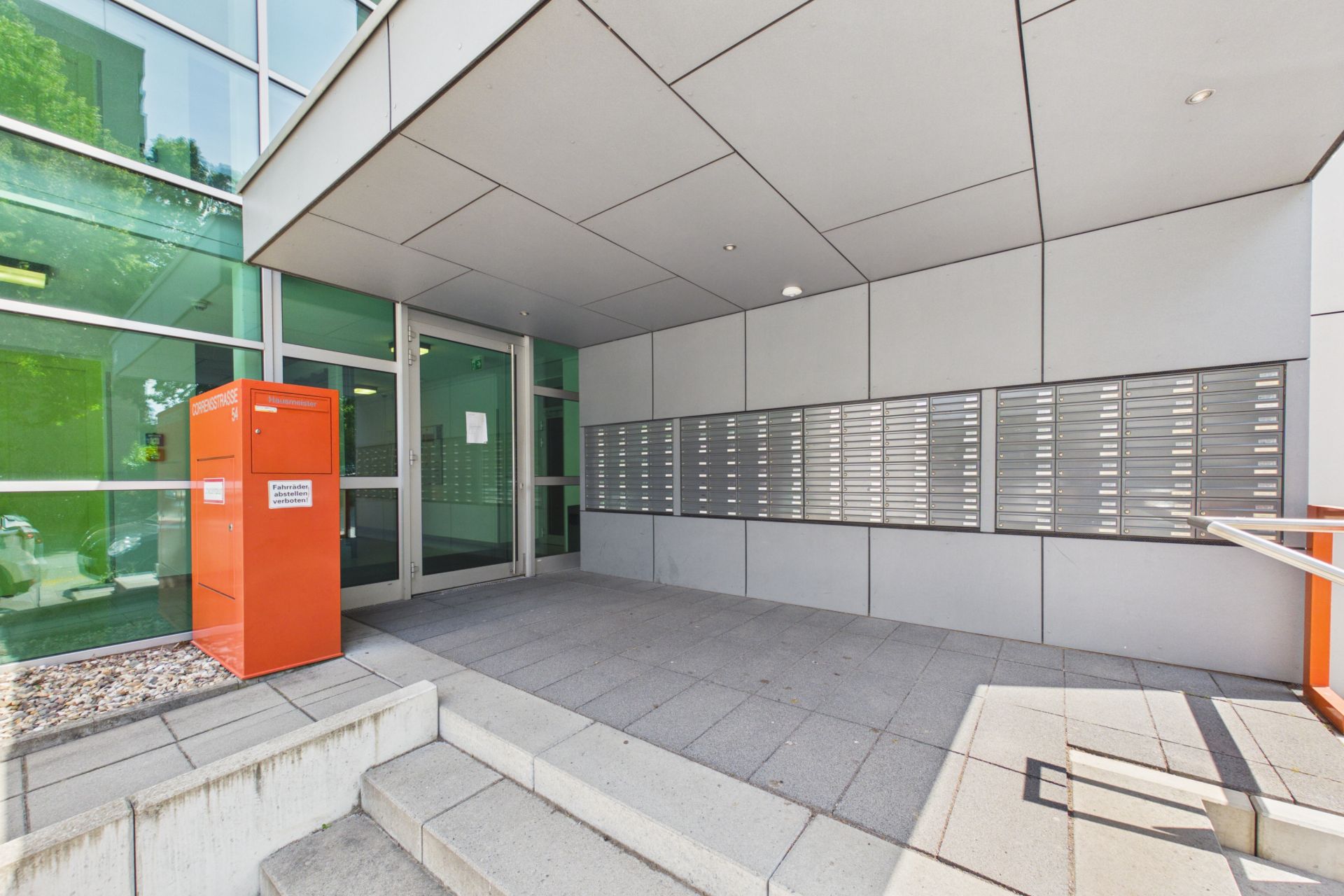 Eingangsbereich eines Wohngebäudes mit Briefkästen und einem orangefarbenen Paketkasten.