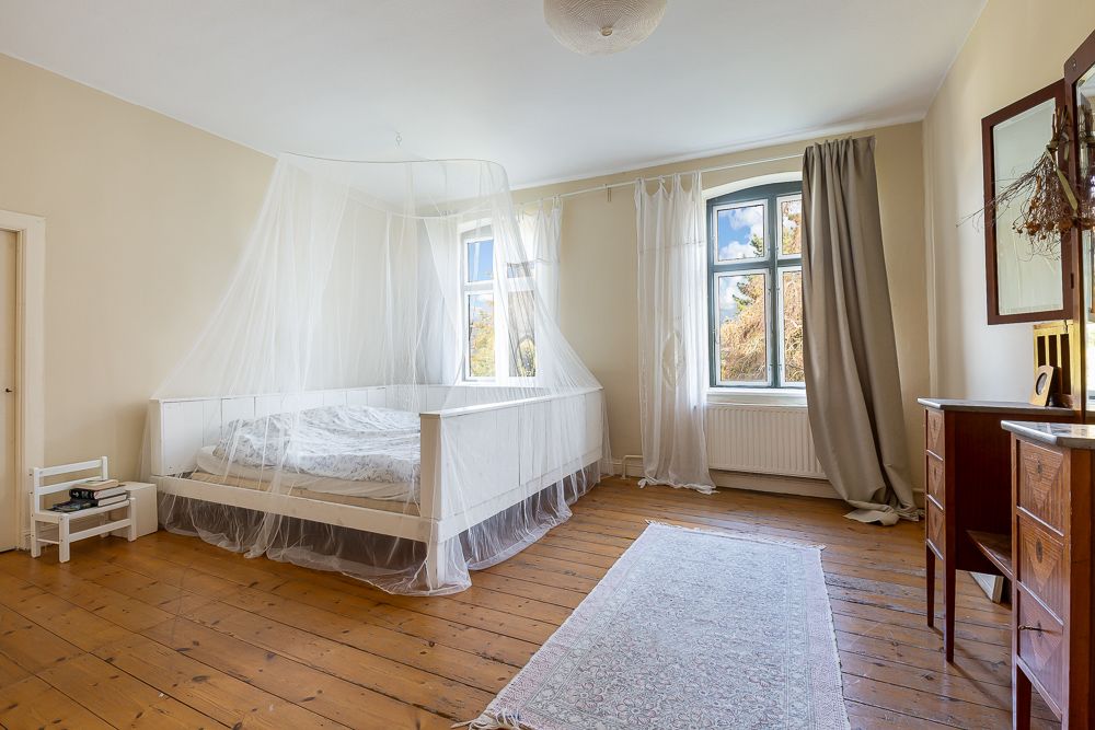 Helles Schlafzimmer mit Holzboden, Himmelbett mit Moskitonetz, Teppich und zwei Fenstern.