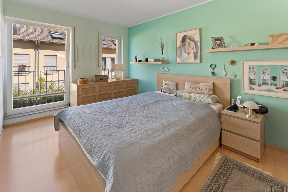 Helles Schlafzimmer mit Doppelbett, Nachttisch, Kommode und Dekoration an türkisfarbener Wand.