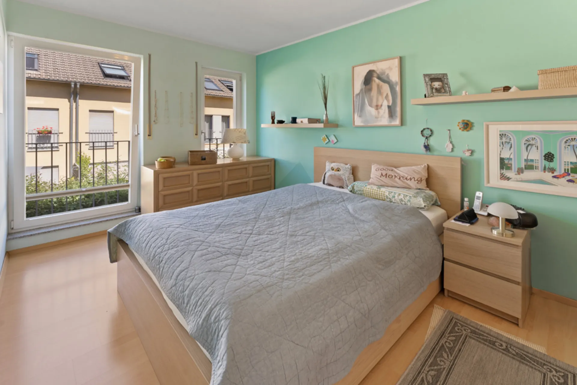 Helles Schlafzimmer mit Doppelbett, Nachttisch, Kommode und Dekoration an türkisfarbener Wand.