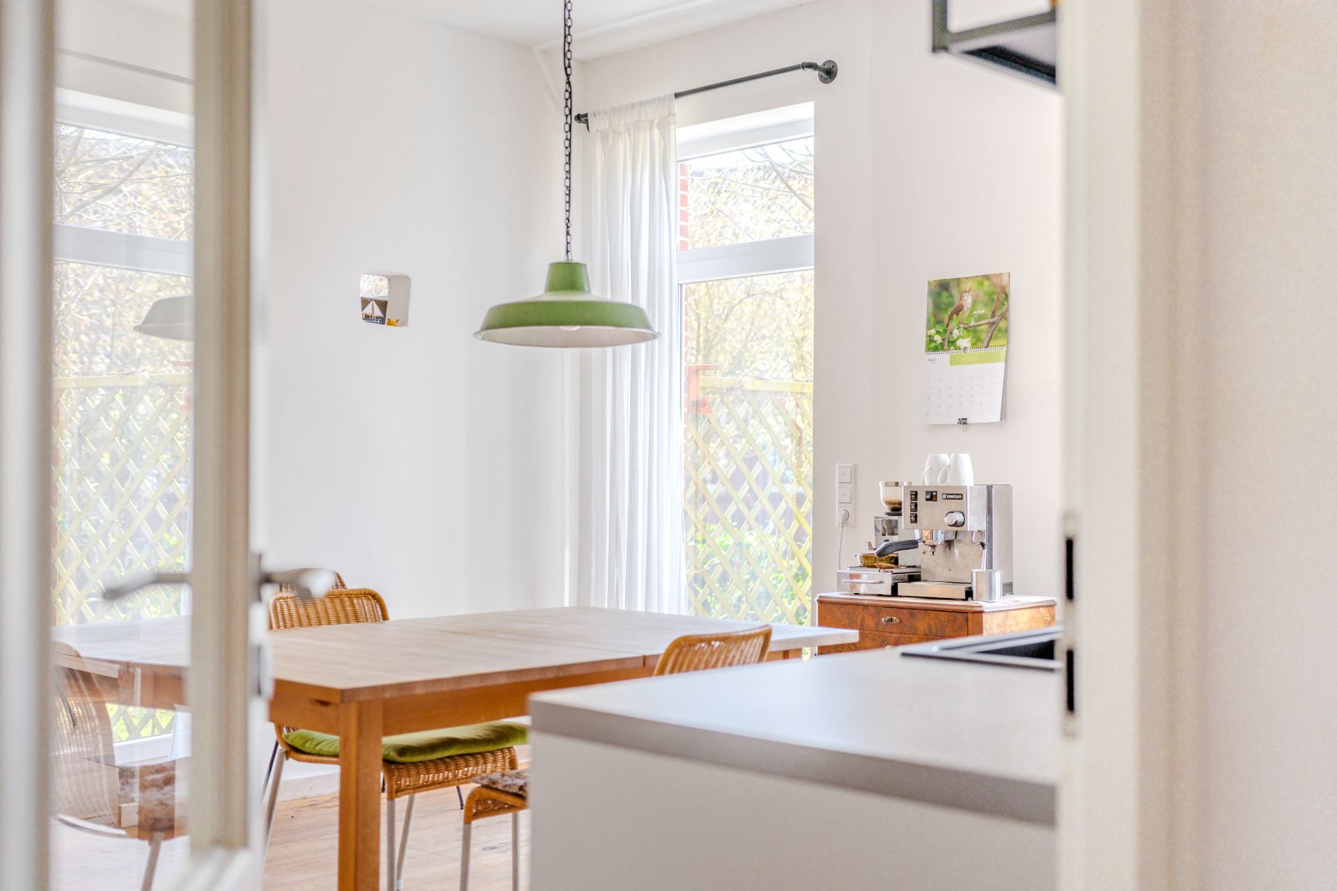 Heller Essbereich mit Holztisch, Stühlen, grüner Hängelampe und großer Fensterfront.