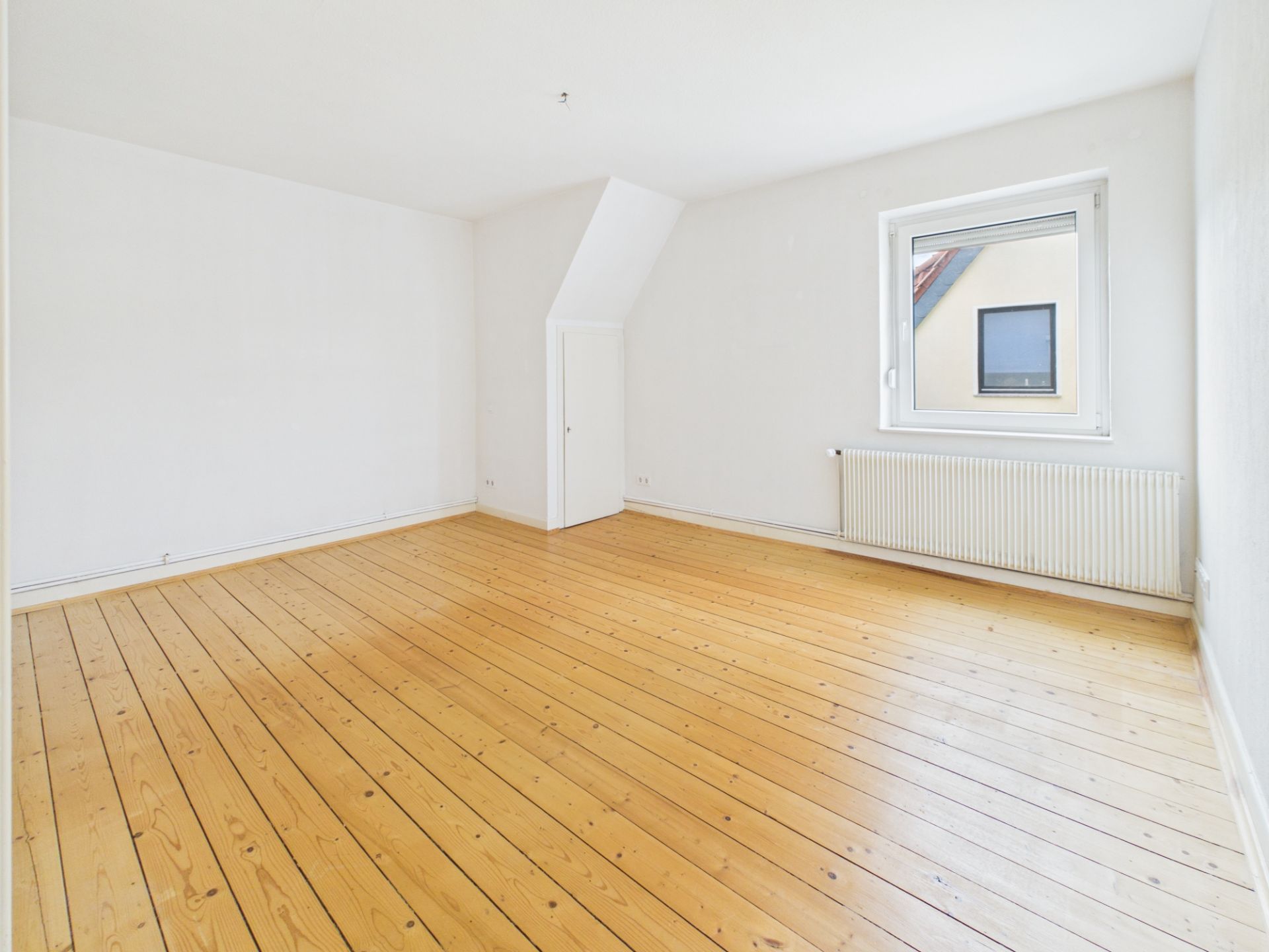 Helles, leeres Zimmer mit Holzboden, weißer Wand, Fenster und Heizung.