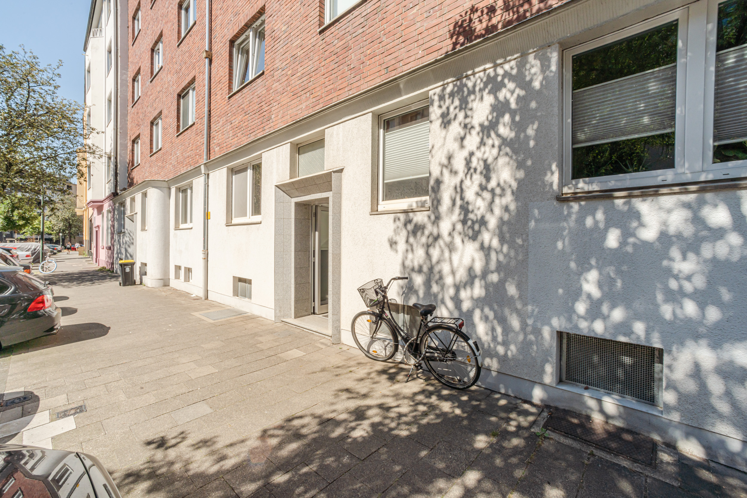 Eingang eines Wohnhauses mit Fahrrad an der Wand, Autos und Bäumen auf dem Gehweg.