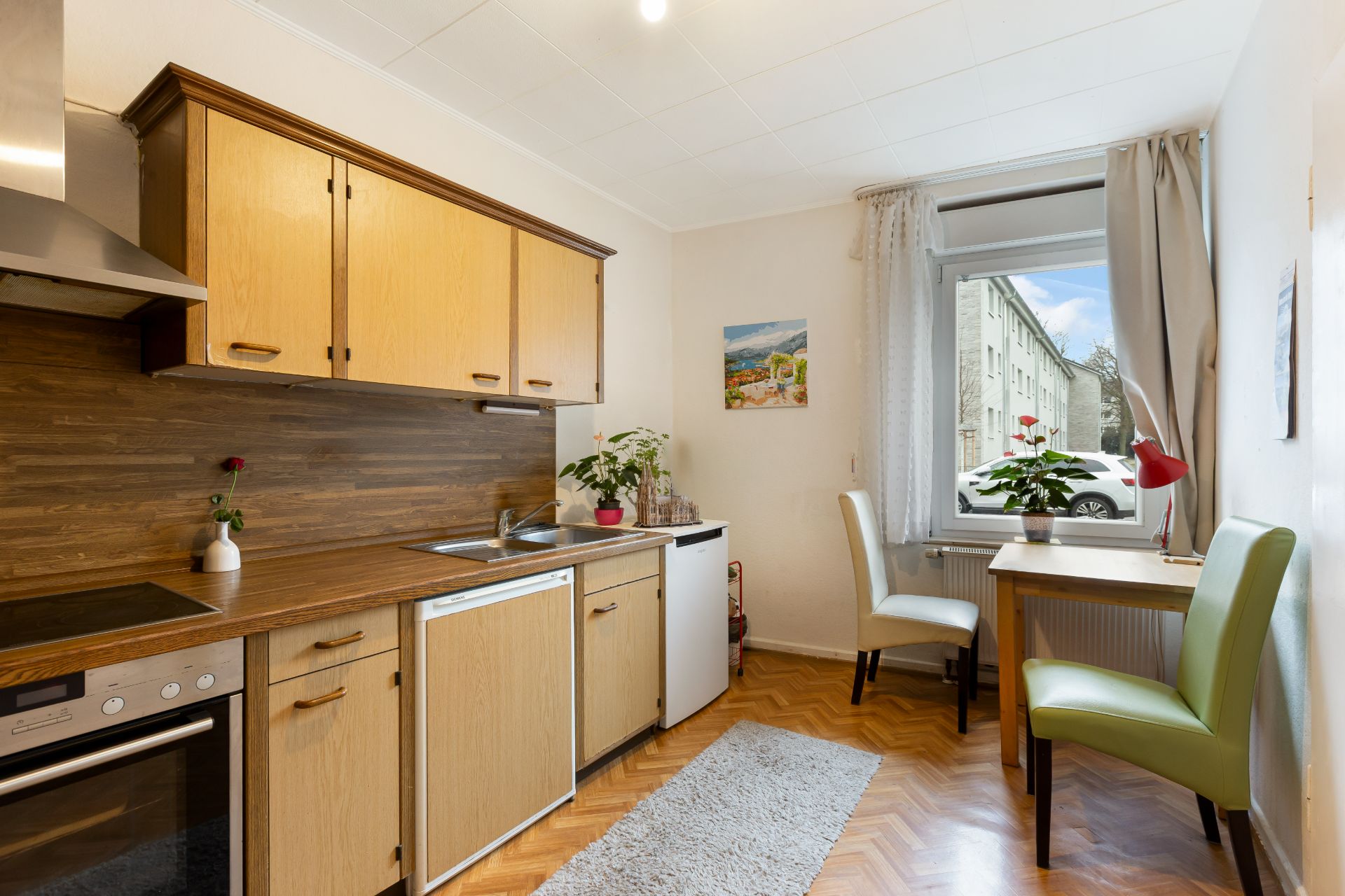 Helle Küche mit Holzmöbeln, kleinem Esstisch, zwei Stühlen und Fenster mit Blick nach draußen.