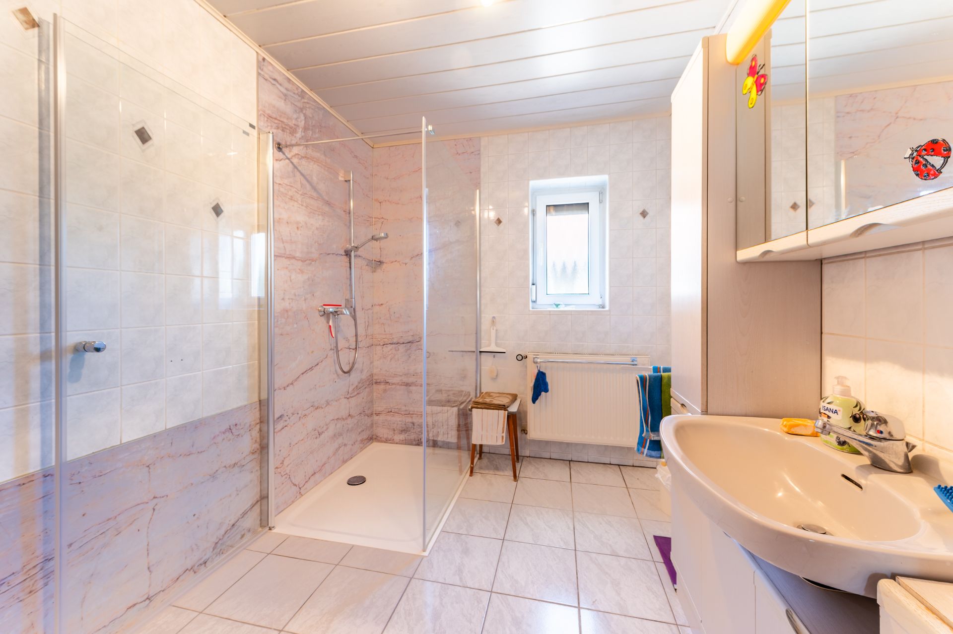 Helles Badezimmer mit ebenerdiger Dusche, Waschbecken, Spiegel und kleinem Fenster.
