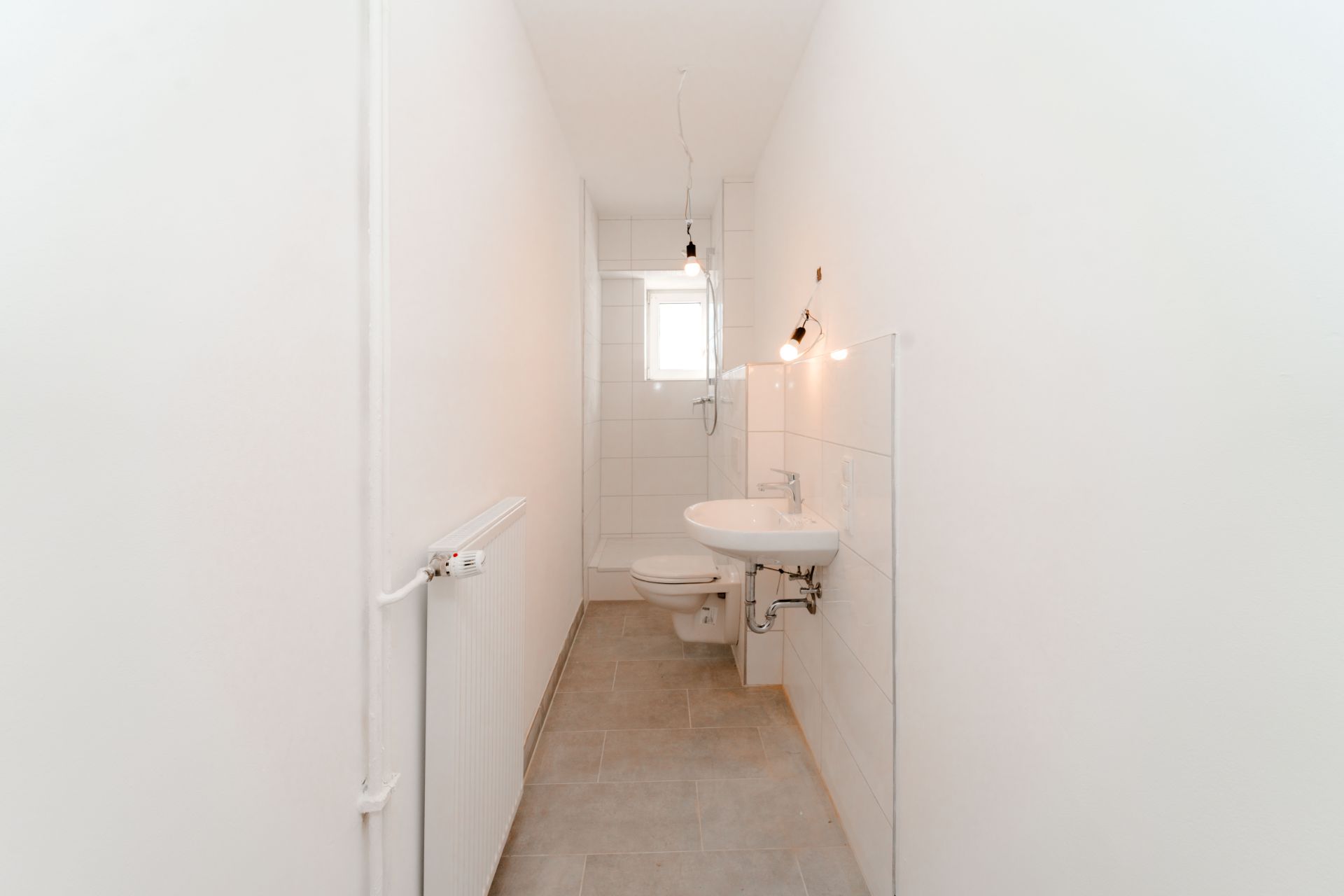 Schmales, weiß gefliestes Badezimmer mit Waschbecken, Toilette und kleinem Fenster.