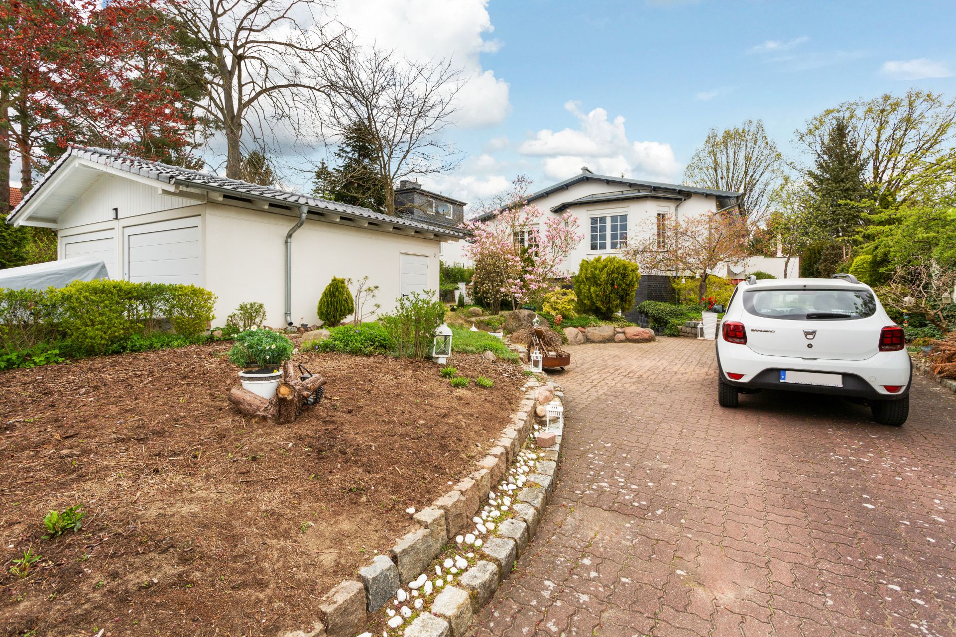 Gepflasterte Einfahrt mit weißem Auto, Garage und Wohnhaus, umgeben von Garten und Bäumen.