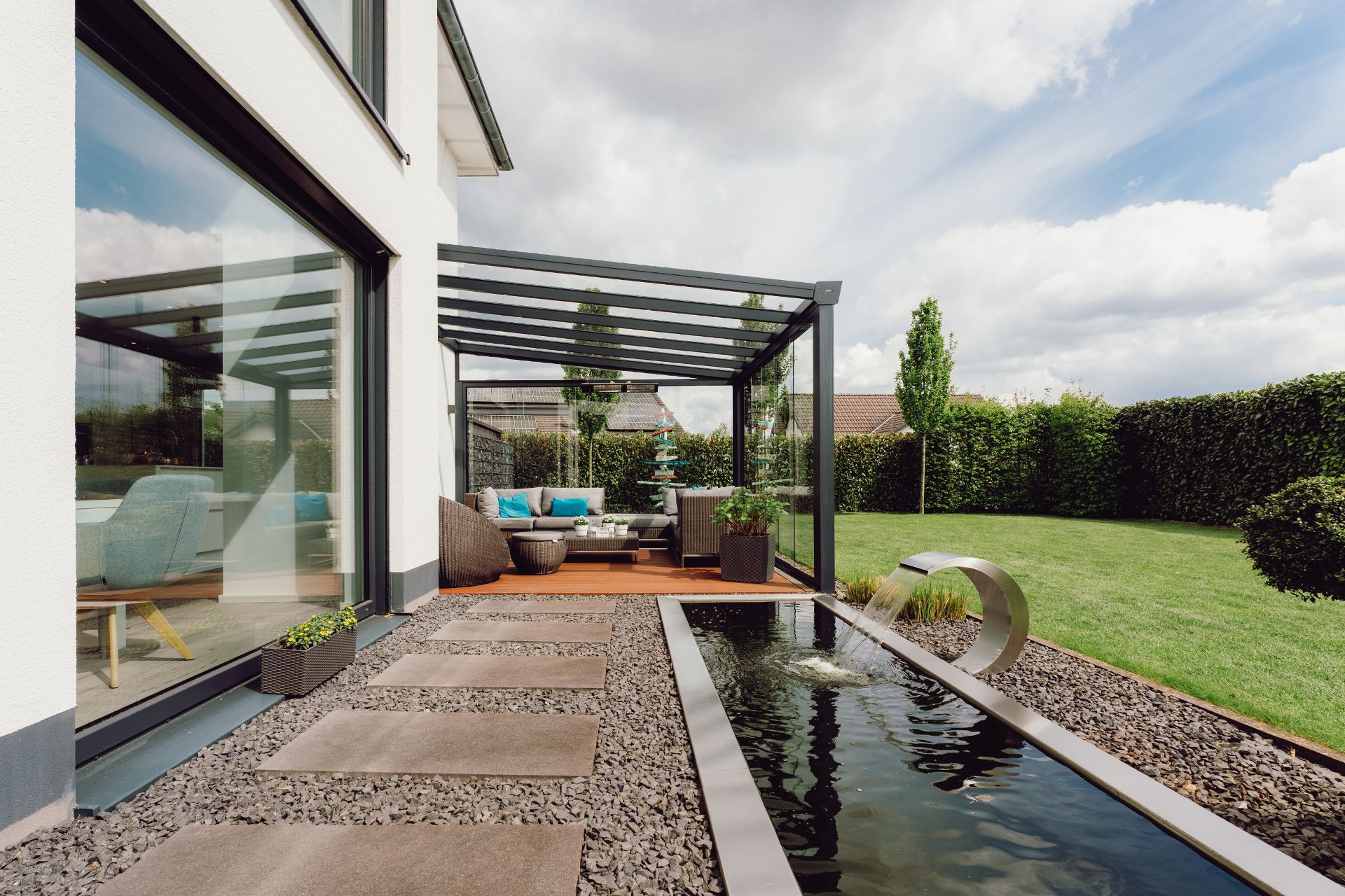 Moderne Terrasse mit Glasüberdachung, Sitzbereich, Teich und gepflegtem Garten.