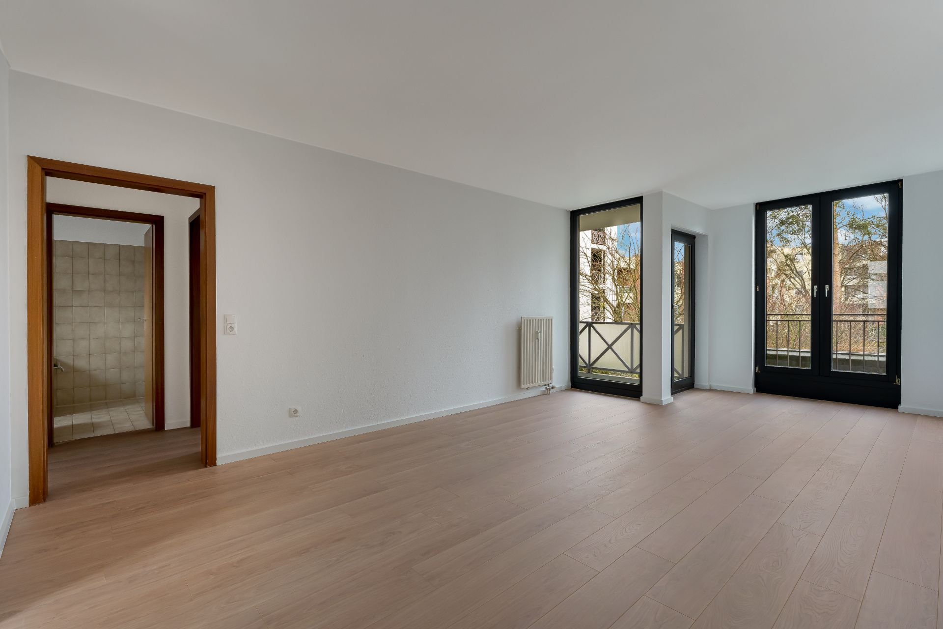 Helles Wohnzimmer mit Holzfußboden, großem Fenster und Zugang zu einem Balkon.