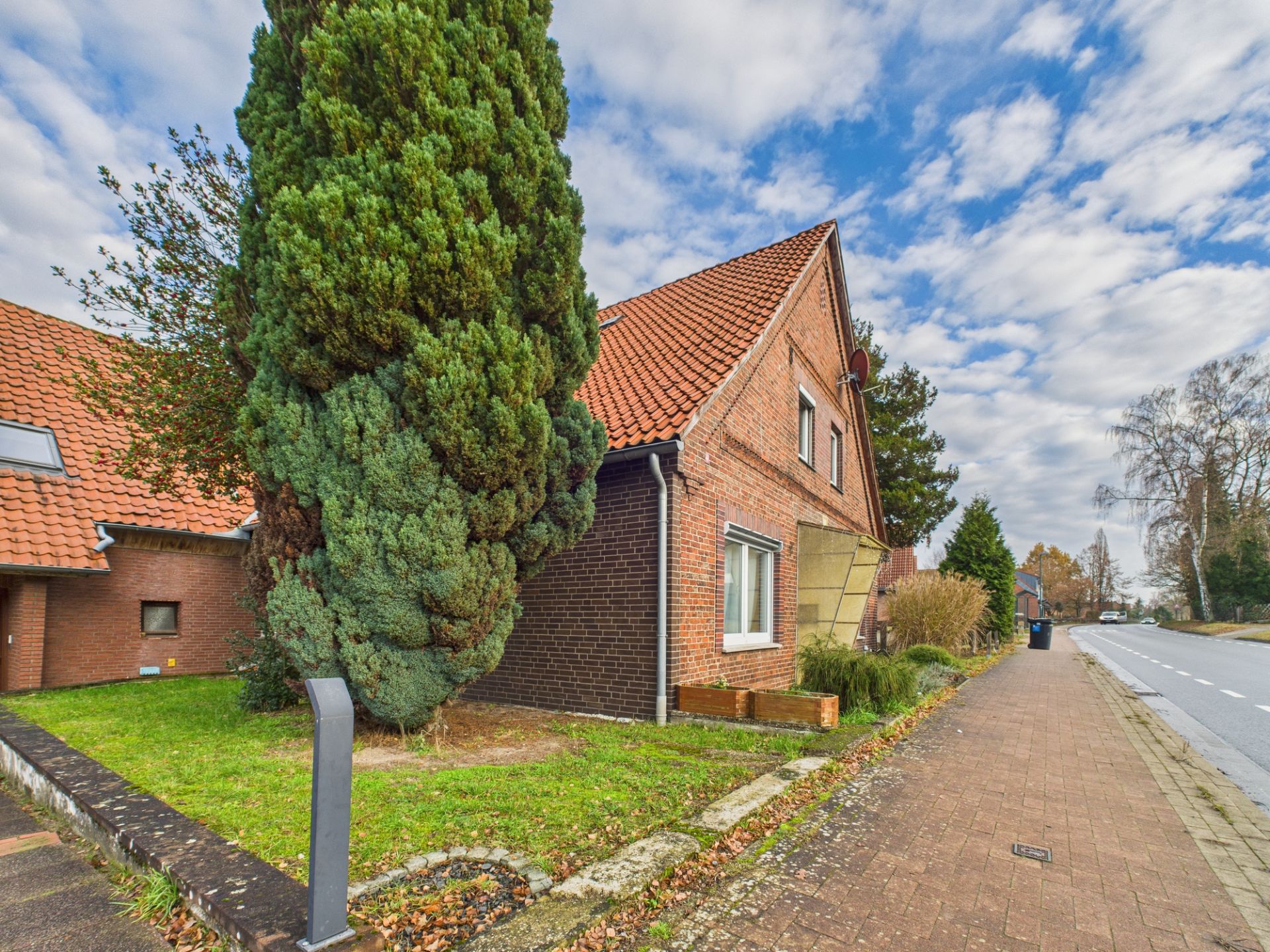 Backsteinhaus mit rotem Dach und großem Baum an einer Straße bei bewölktem Himmel.