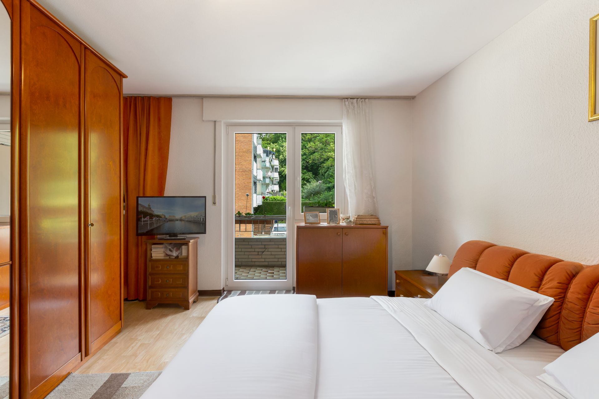 Helles Schlafzimmer mit Doppelbett, Schrank, TV und Zugang zu einem Balkon.