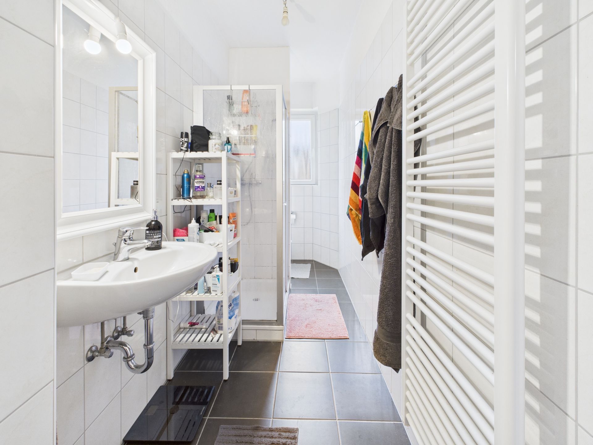 Helles Badezimmer mit Waschbecken, Regal, Dusche, Handtüchern und Heizkörper.