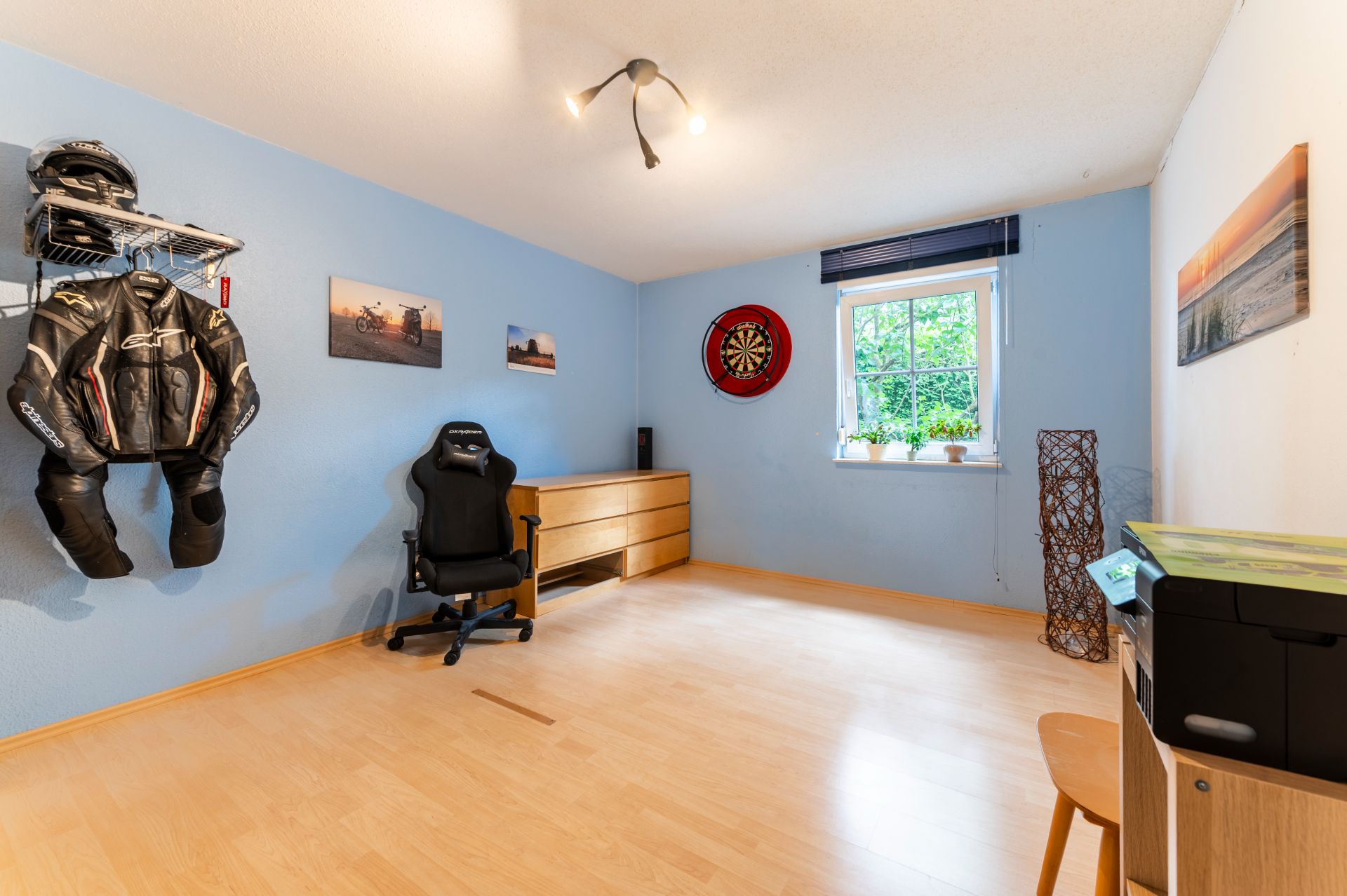Helles Zimmer mit Holzfußboden, Schreibtischstuhl, Kommode, Dartscheibe und Fenster zur Außenseite.
