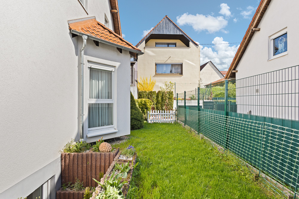 Schmaler Gartenbereich mit Rasen, Blumenbeet und Zaun zwischen zwei Wohnhäusern.