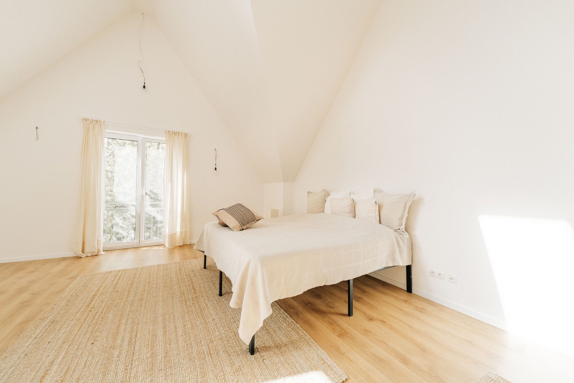 Helles Schlafzimmer mit Doppelbett, Holzboden und großem Fenster mit Vorhängen.