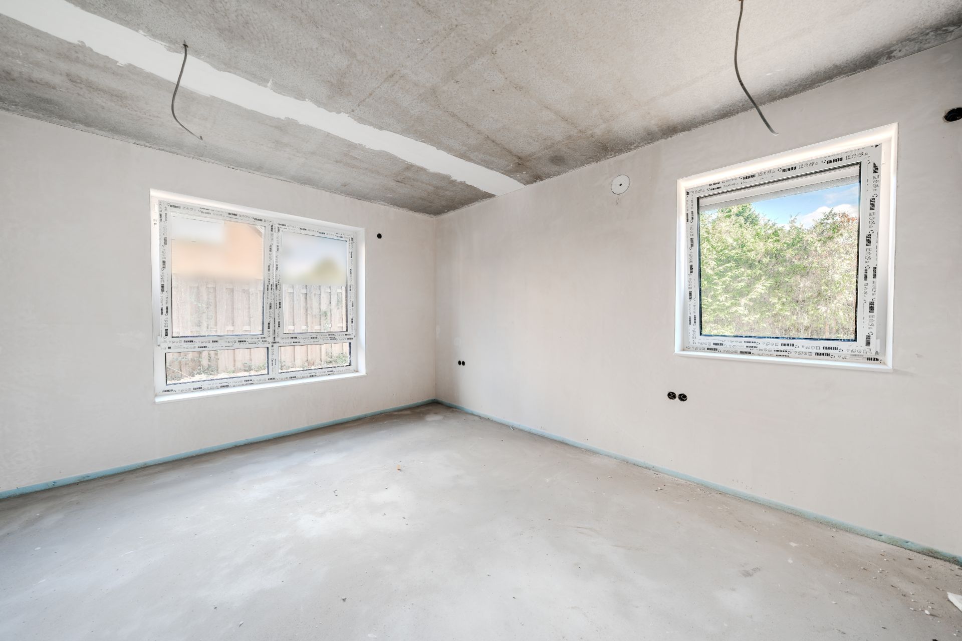 Leerer, unfertiger Raum mit zwei Fenstern und sichtbaren Kabeln an der Decke.