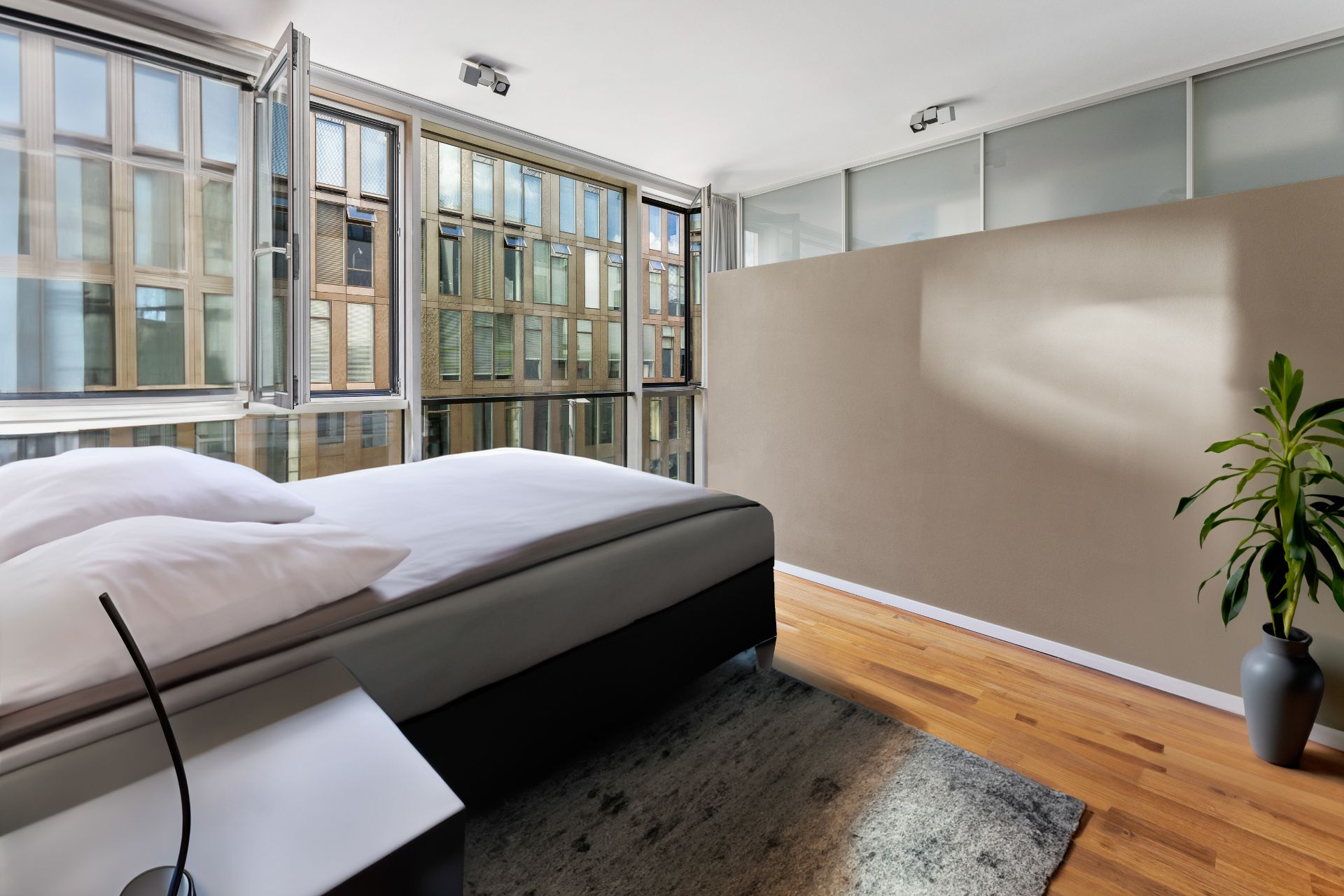 Modernes Schlafzimmer mit großem Fenster, Bett, Nachttisch, Teppich und Pflanze auf Holzfußboden.
