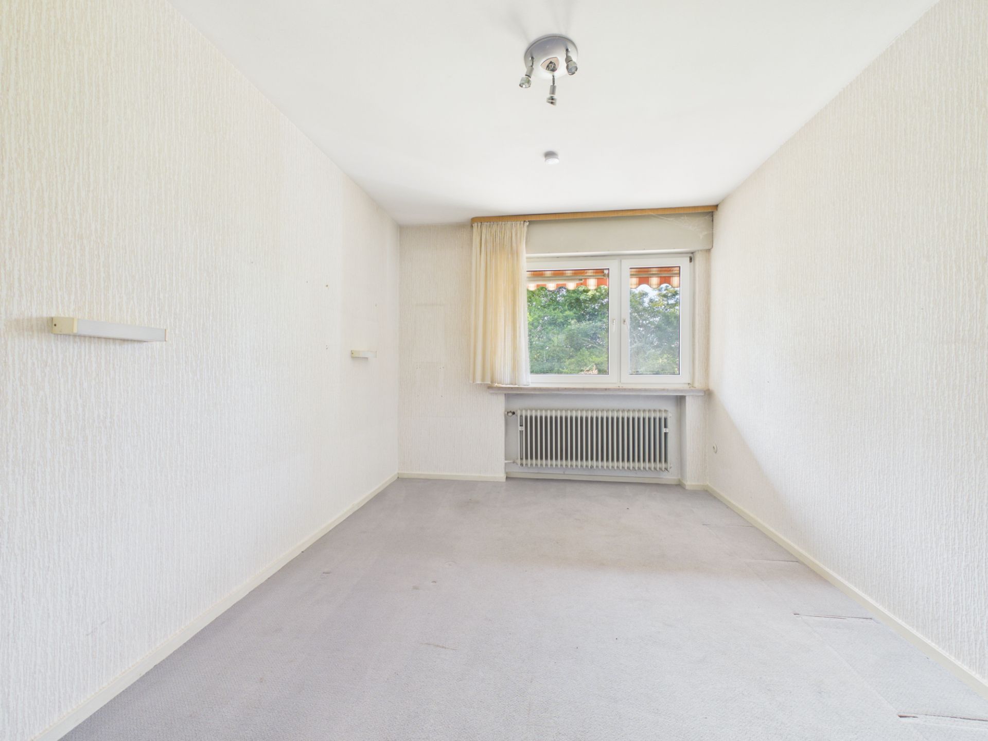 Heller, leerer Raum mit Teppichboden, Fenster und Heizung unter dem Fenster.
