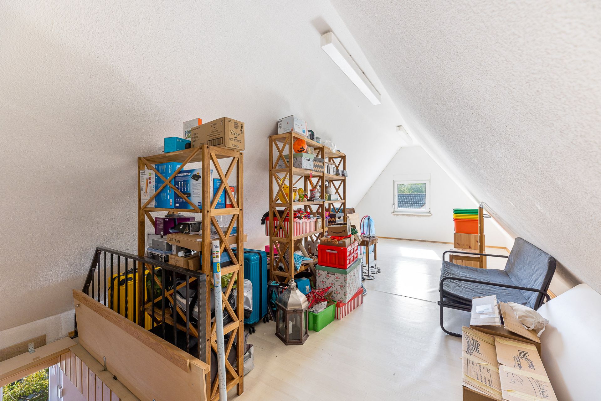 Dachzimmer mit Regalen, Kisten und einem kleinen Fenster, helle Wände und ein Sofa.