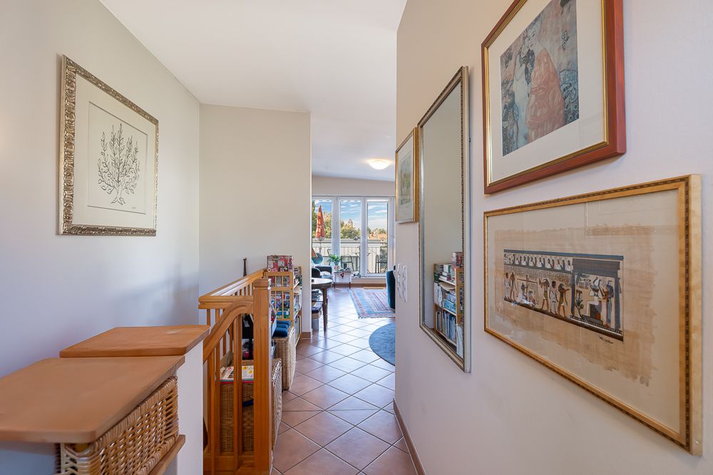 Heller Flur mit Bildern an der Wand, Holzgeländer und Blick in ein Wohnzimmer mit großen Fenstern.