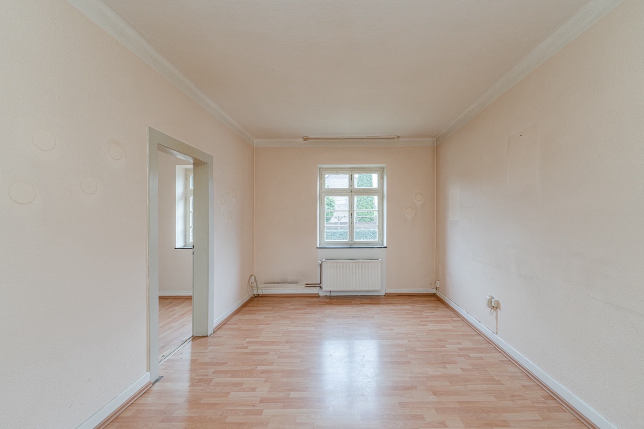 Leerer, heller Raum mit Holzfußboden, Fenster und Türöffnung zu einem weiteren Zimmer.