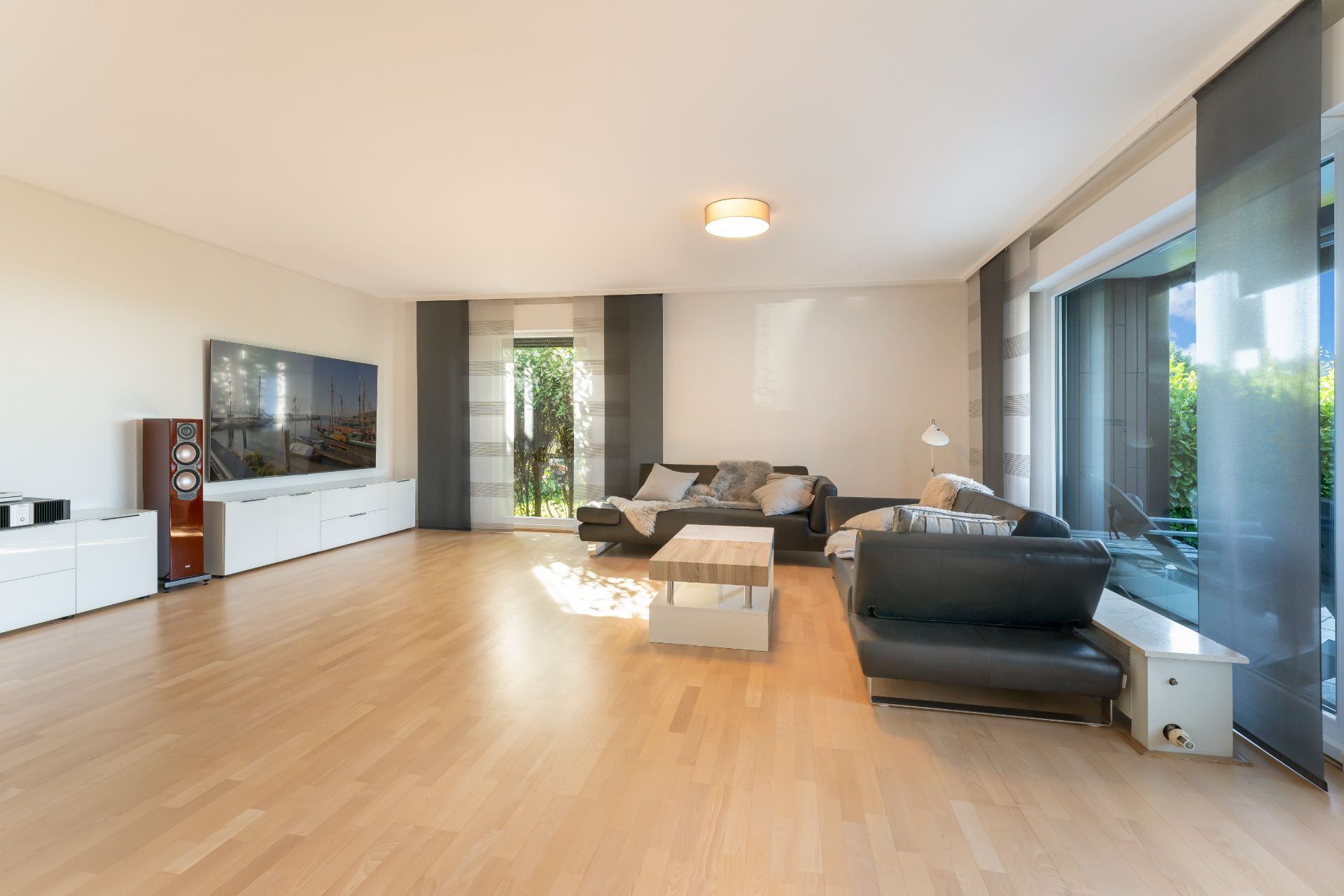 Modernes Wohnzimmer mit Holzfußboden, großen Fenstern, zwei Sofas und einem Fernseher.