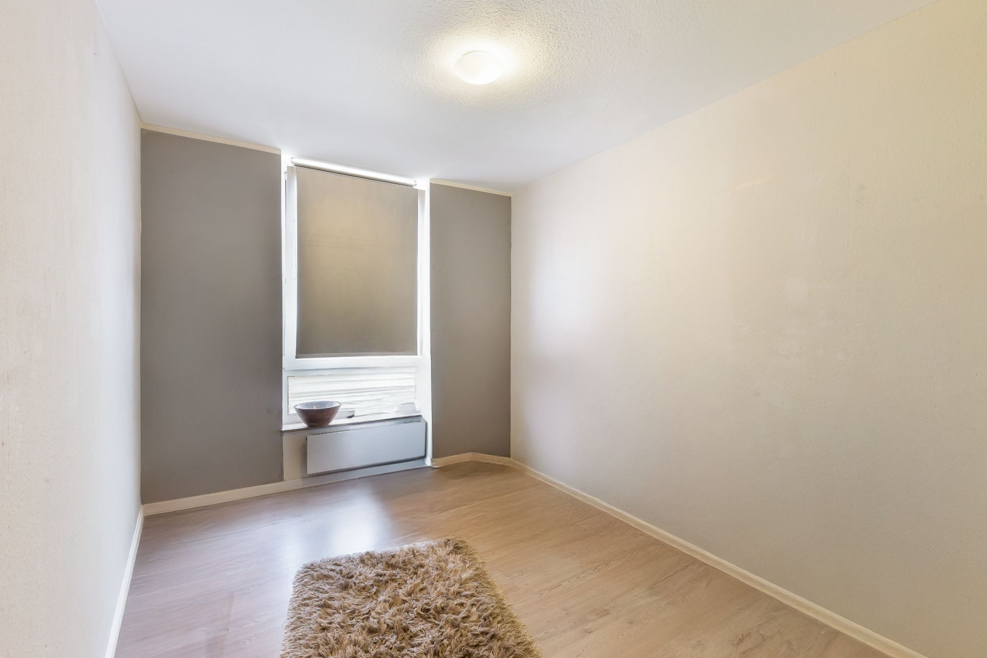 Leeres Zimmer mit Holzfußboden, Fenster mit Jalousie und kleinem Teppich auf dem Boden.