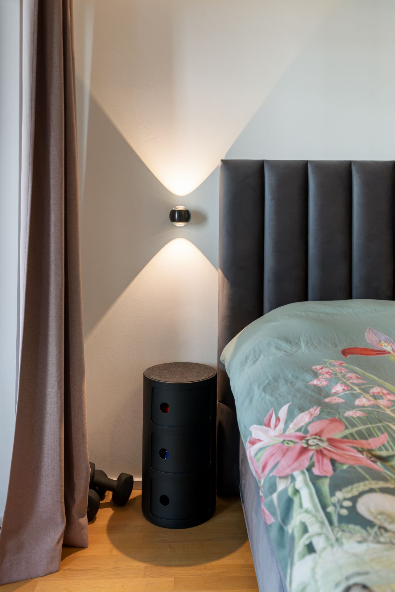 Bett mit Blumen-Bettwäsche, schwarze Kommode, Wandlampe und Hanteln auf Holzfußboden.