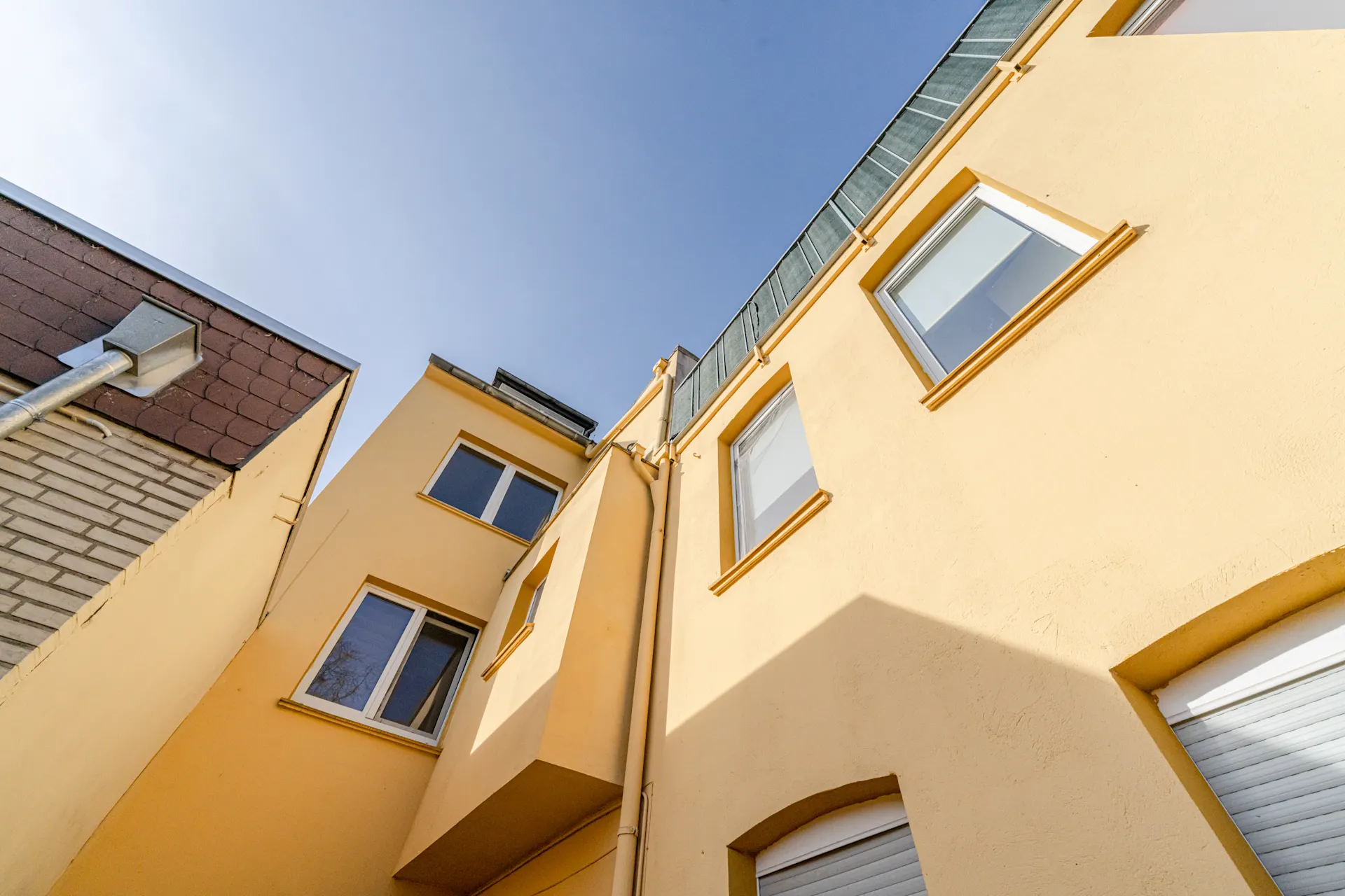 Blick nach oben auf gelbe Wohnhausfassade mit mehreren Fenstern bei blauem Himmel.
