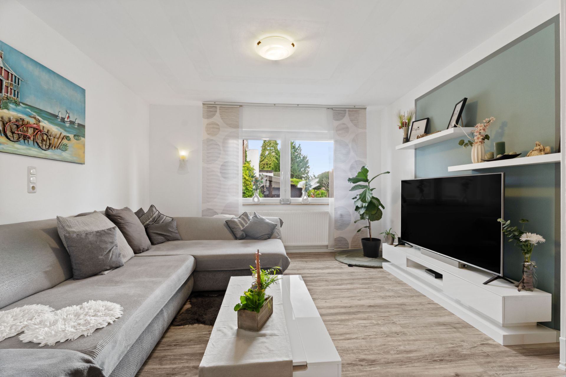 Modernes Wohnzimmer mit grauem Sofa, Couchtisch, Fernseher und großem Fenster mit Blick ins Grüne.