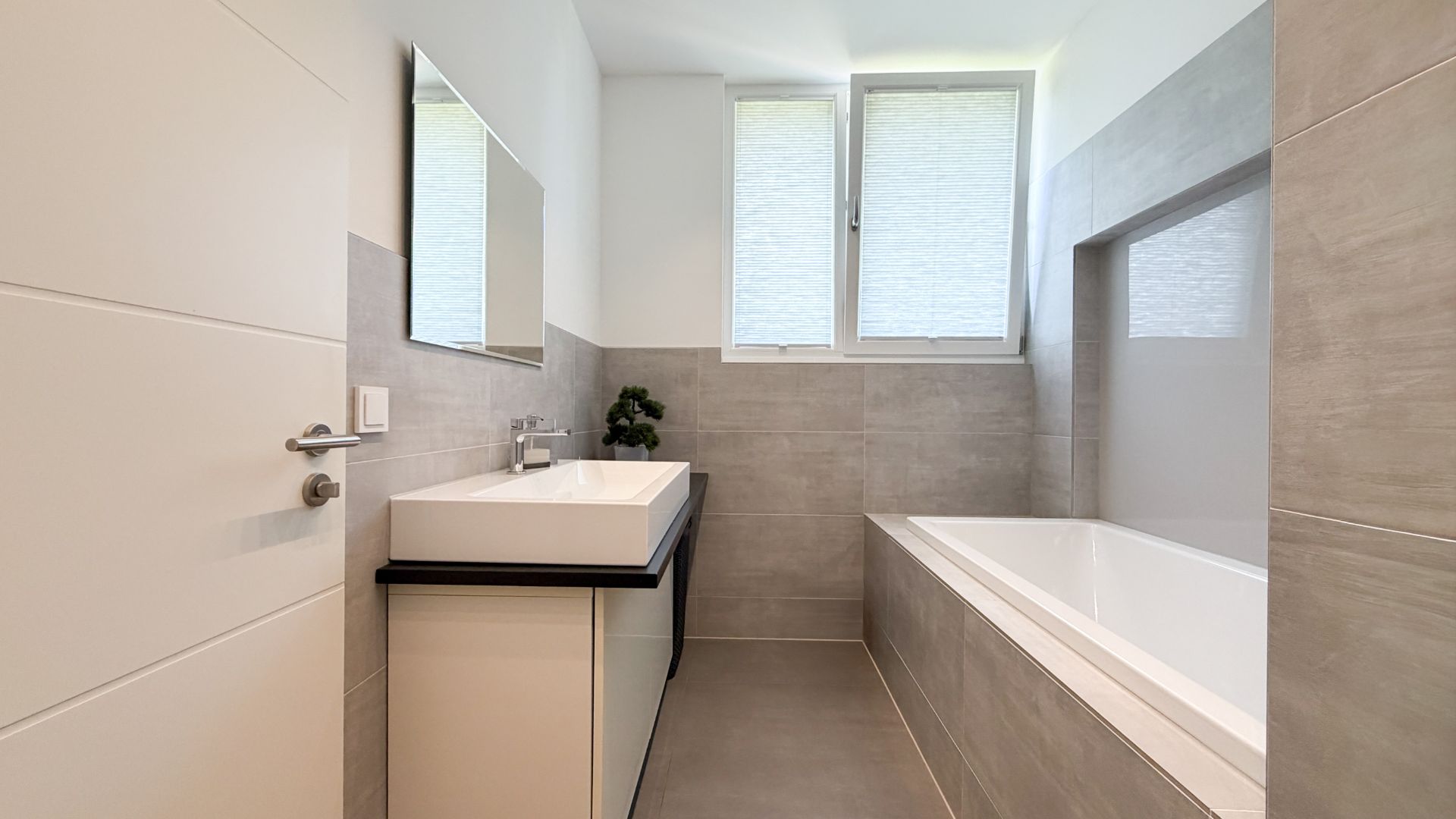 Modernes Badezimmer mit Waschbecken, Spiegel, Badewanne und zwei Fenstern mit Plissees.
