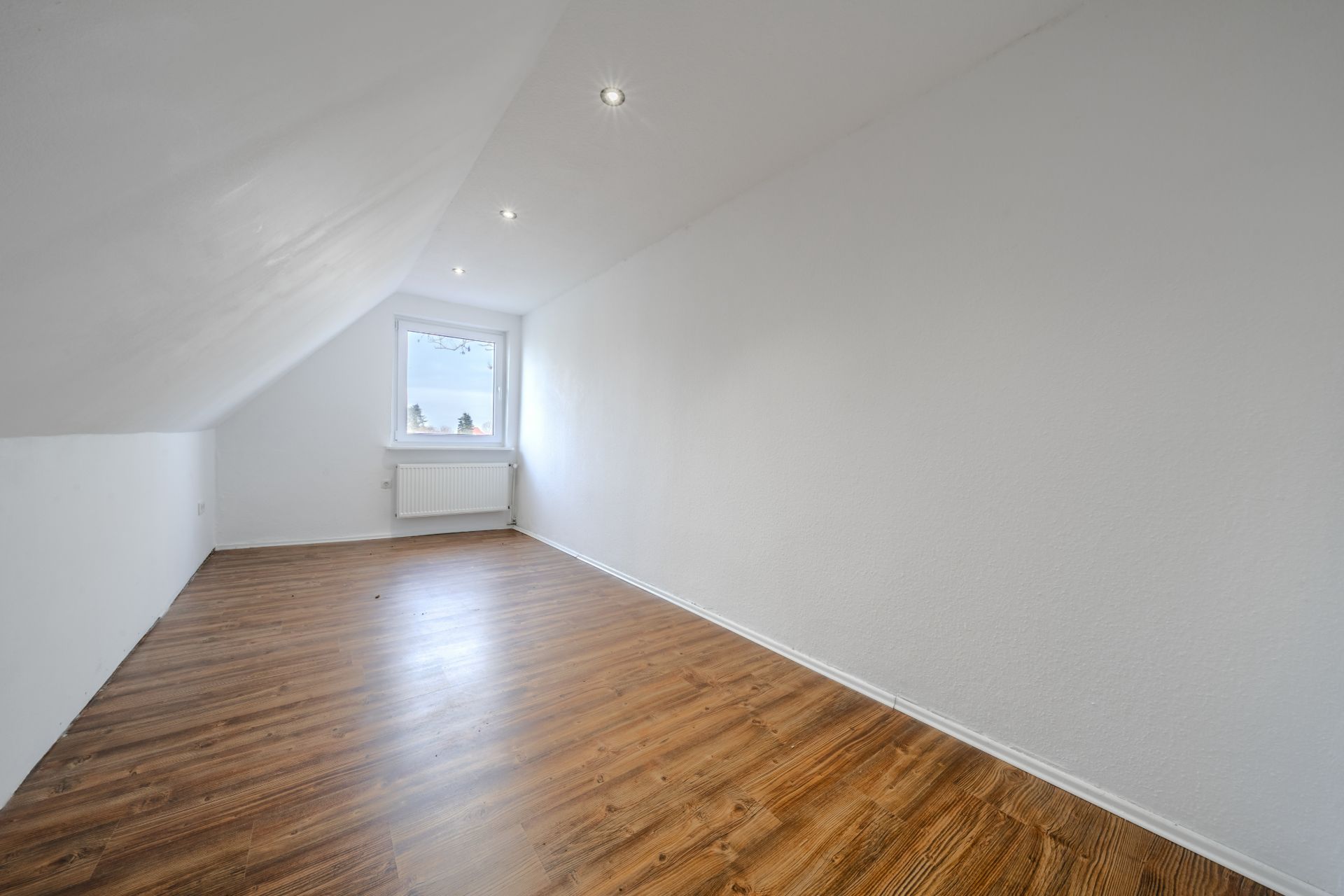 Leeres Zimmer mit schräger Decke, weiß gestrichenen Wänden und Holzfußboden, Fenster am Ende.