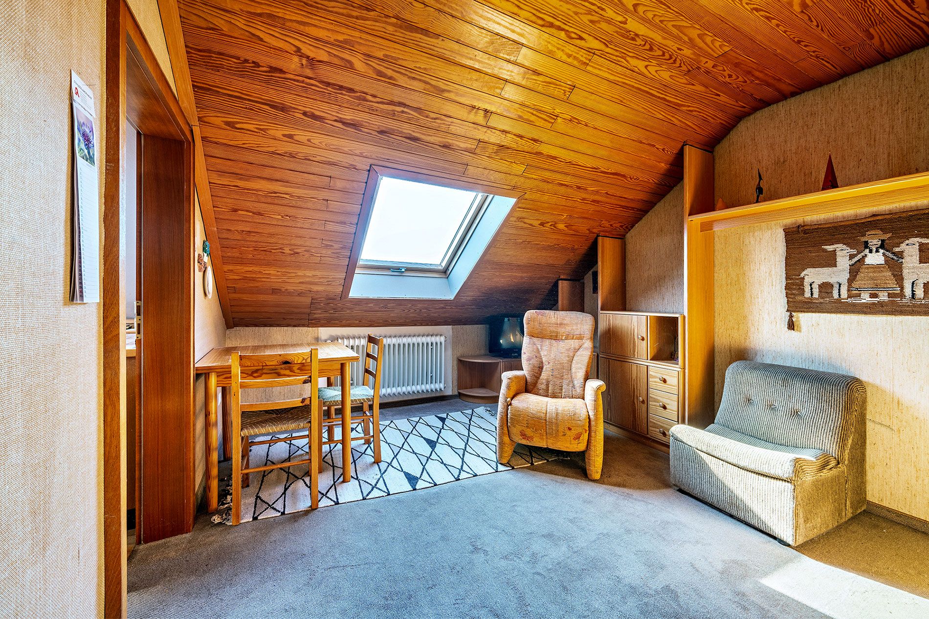 Wohnzimmer mit holzverkleideter Dachschräge, Sessel, Stuhlgruppe, Teppich und Dachfenster.