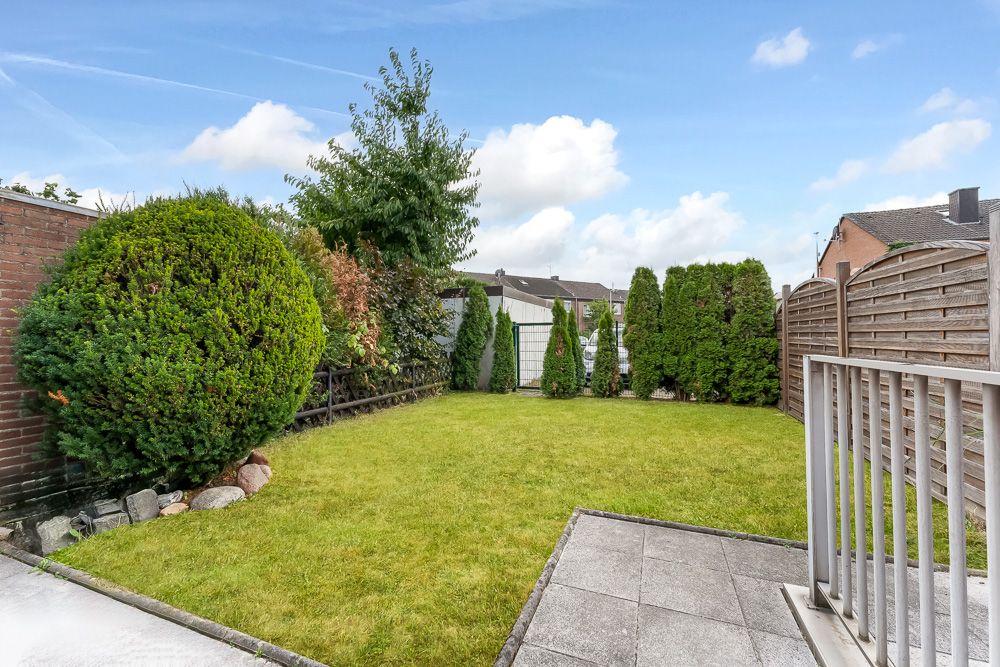 Kleiner Garten mit Rasenfläche, Hecken, Baum und Terrasse, umzäunt von Zäunen und Mauer.