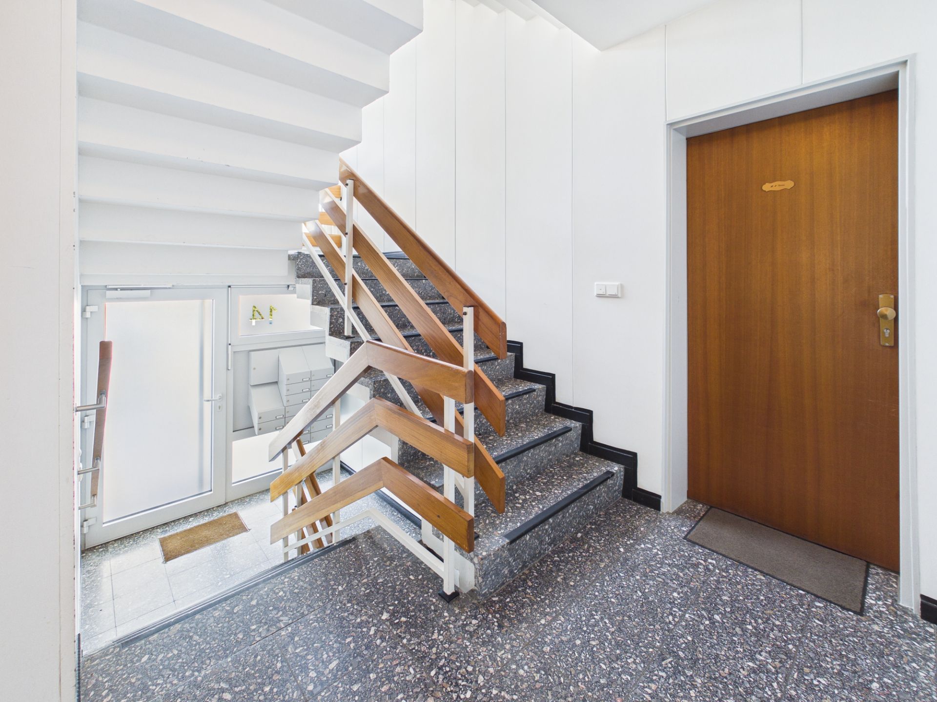 Treppenhaus mit grauem Boden, Holzgeländer und brauner Wohnungstür.