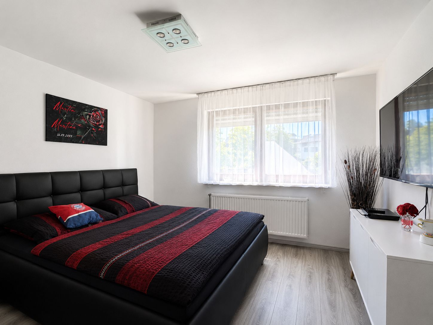 Modernes Schlafzimmer mit großem Bett, Wandbild, Fenster mit Gardinen und TV an der Wand.