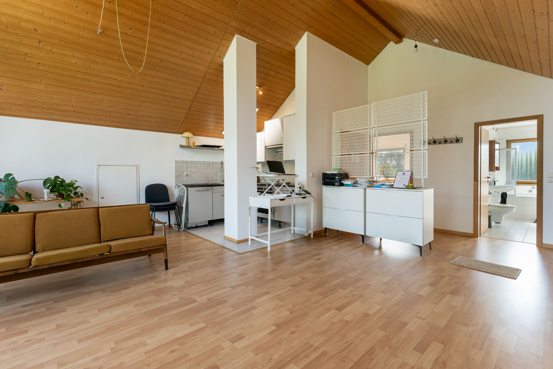 Helles Wohnzimmer mit Holzdecke, Parkettboden, Sofa, Schreibtisch und offener Küche.