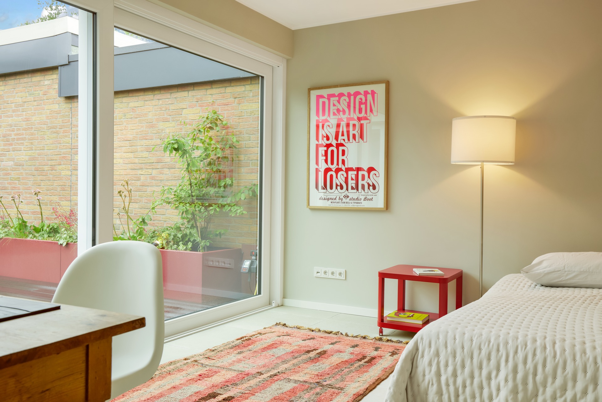 Helles Schlafzimmer mit großem Fenster, Stehlampe, rotem Beistelltisch und Wandbild.