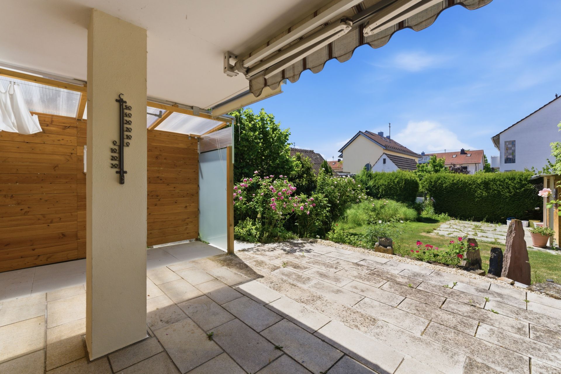 Überdachte Terrasse mit Fliesenboden, Sicht auf Garten mit Blumen und Nachbarhäuser.